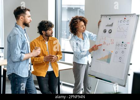 Un team di colleghi multirazziali motivati e di successo che lavorano nella stessa azienda, si trova in ufficio vicino alla lavagna con grafici finanziari, analizza il concetto e la strategia di un progetto Foto Stock