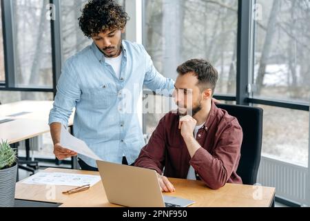 Due uomini di etnia indiana e caucasica, lavorano colleghi, lavorano insieme su un progetto in un ufficio moderno, si consigliano, condividono idee e soluzioni, progettano una strategia di promozione del prodotto Foto Stock