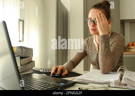 una giovane donna bianca lavora a distanza da casa, ci sono un sacco di cartelle con documenti sul tavolo, è molto stanca, la sua testa fa male Foto Stock