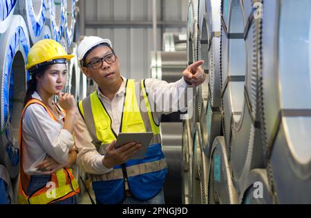 Il Senior Engineer Manager forma i nuovi dipendenti all'interno della fabbrica di lamiere. Tutti indossano giubbotto di sicurezza e elmetto. La pila di rulli in lamiera di acciaio si trova in Foto Stock
