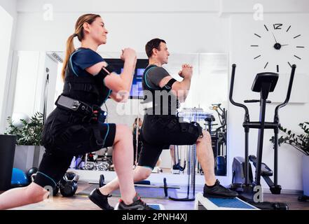 La coppia sportiva in abiti ems è impegnata in attività fisica in fitness club o palestra. Uomo e donna con mani stringate che fanno affondi in avanti con STEP pla Foto Stock