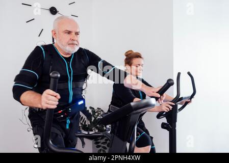 Due persone anziane in abiti ems sono impegnate in attrezzature sportive. L'uomo anziano serio sta esercitandosi sull'addestratore ellittico. Donna anziana che pedala exerci Foto Stock