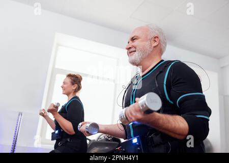 Coppia sorridente più anziana in vestiti di ems che fanno il workout elettrico di stimolazione del muscolo con i dumbbells in palestra o nel randello di idoneità. Uomo dai capelli grigi e biondo a pelo w Foto Stock