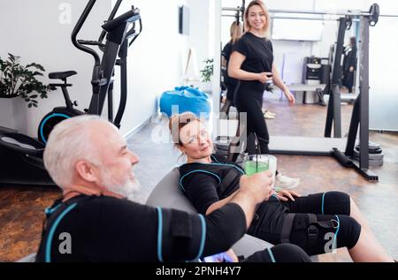 Uomo sorridente e donna bionda di mezza età in abiti ems bere scuoti sani seduti in sedie da massaggio. Coppia di occhiali da vista e riposo Foto Stock