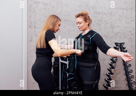 Allenatore femminile si mette in tuta per l'allenamento ems su donne anziane. Il formatore fissa i fili sul torace del cliente. Preparazione per la stimolazione elettrica del muscolo t Foto Stock