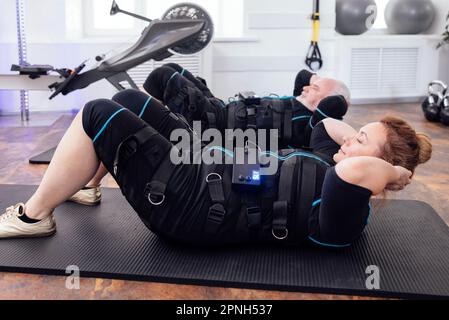 Coppie sportive di anziani in abiti ems si esercitano in palestra o club fitness. Uomo grigioso e donna grumo che pompano i muscoli addominali sul pavimento Foto Stock