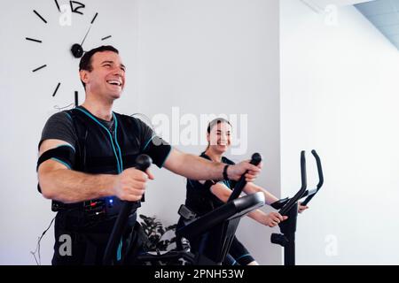 Un paio di sportivi in abiti ems sono impegnati in attrezzature sportive nel fitness club o palestra. Ridendo giovane uomo sta esercitandosi sull'addestratore ellittico. P Foto Stock