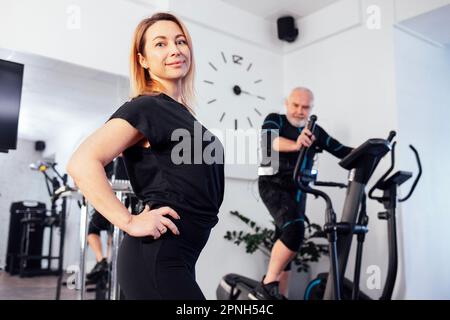 Personal trainer femminile con il suo cliente in abito ems in palestra o sala fitness. Sport l'uomo anziano grayhaired sta esercitandosi sull'addestratore ellittico. Elettr Foto Stock