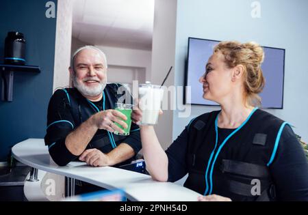 Uomo e donna sorridenti senior in abiti ems frullati da bibita nel bar dello sport. Le coppie anziane comunicano e riposano dall'addestramento con lo stimolo elettrico del muscolo Foto Stock