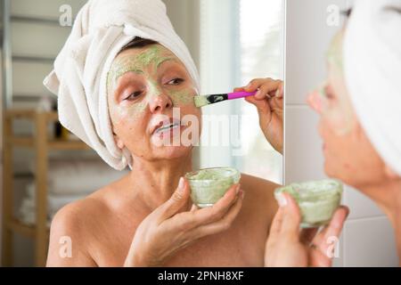 Matura bella donna che applica verde fatto in casa maschera facciale fai-da-te sulla pelle. Guardavano nello specchio del bagno, avvolti in un asciugamano. Routine per la cura della pelle Foto Stock