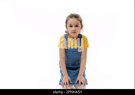 Bambina caucasica in t-shirt gialla e denim sundress tenendo le mani sulle ginocchia mentre si siede su sfondo bianco Foto Stock