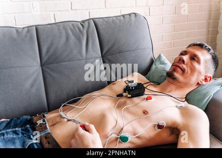 Uomo sdraiato su un divano con un cardiofrequenzimetro Holter collegato Foto Stock
