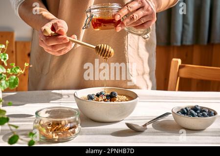 Donna che prepara sana dieta vegana colazione nutriente. Femmina a mano che versa il miele nella ciotola con porridge di farina d'avena con noci e mirtilli. Foto Stock