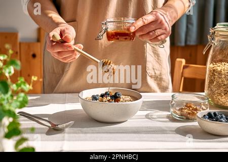 Donna che prepara sana dieta vegana colazione nutriente. Femmina a mano che versa il miele nella ciotola con porridge di farina d'avena con noci e mirtilli. Foto Stock