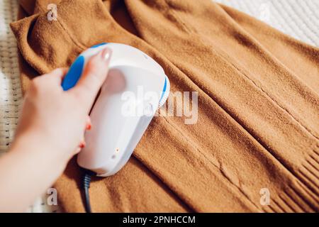 Donna che rimuove il pelucchi dai vestiti usando il rasoio fuzz a casa. Primo piano di un dispositivo elettrico portatile cablato. Cura degli indumenti Foto Stock