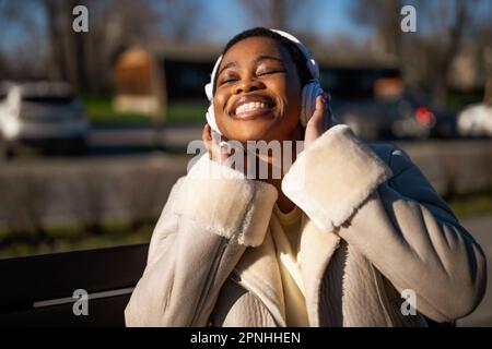 Ritratto all'aperto di felice donna nera nella giornata di sole. Sta ascoltando musica con le cuffie e rilassandosi. Foto Stock