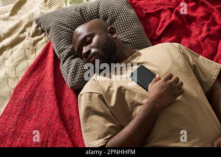 Vista dall'alto dell'uomo afroamericano esausto che dorme a letto con il telefono cellulare Foto Stock