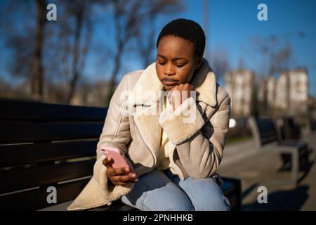 Ritratto all'aperto di una donna nera preoccupata nelle giornate di sole. È seduta su una panchina per strada e la messaggistica al telefono. Foto Stock