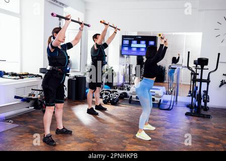 Sport Couple in ems si allenano con personal trainer in fitness club. I giovani tengono i bastoni della palestra nelle mani e fanno squat. Il coach mostra l'e Foto Stock