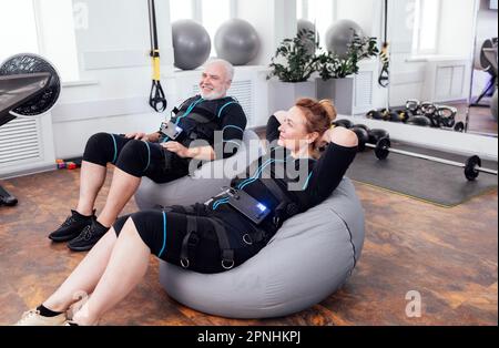 Le coppie anziane si siedono sulle sedie di massaggio mentre aspettano per l'allenamento di ems nella palestra o nel centro fitness. Uomo e donna senior con capelli rossi in abiti ems sempre pronti Foto Stock