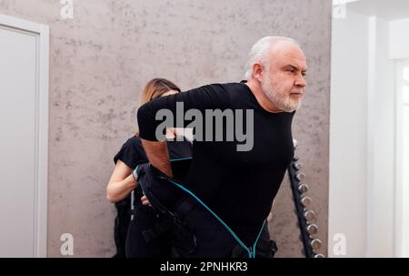 L'istruttore femmina fissa le attrezzature necessarie su ems Suit al cliente maschio. Il coach di idoneità mette sul costume per l'allenamento elettrico di stimolazione del muscolo sul anziano Foto Stock