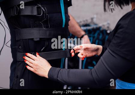 L'istruttore femmina fissa le attrezzature necessarie su ems Suit al cliente maschio. Il coach di idoneità mette sul costume per l'allenamento elettrico di stimolazione del muscolo sul anziano Foto Stock