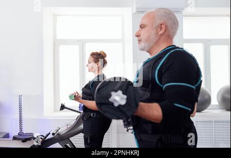 Coppia sorridente più anziana in vestiti di ems che fanno il workout elettrico di stimolazione del muscolo con i dumbbells in palestra o nel randello di idoneità. Uomo dai capelli grigi e biondo a pelo w Foto Stock