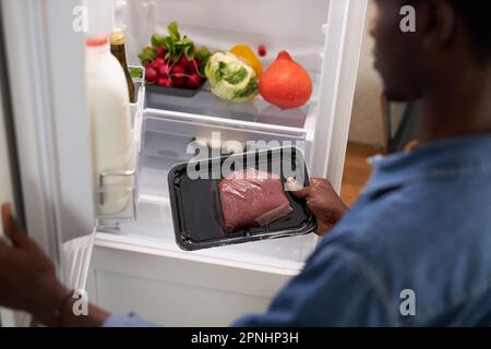 Primo piano di un giovane uomo nero che tollera il manzo dal frigorifero mentre cucinava la cena a casa Foto Stock