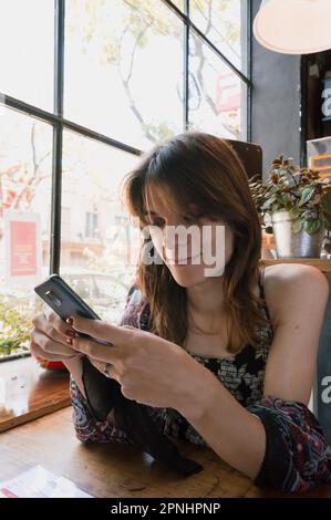 Giovane donna sottile Latina con capelli corti, pelle discreta e etnicità transgender argentina, è sola utilizzando il telefono nel ristorante, seduta in attesa Foto Stock