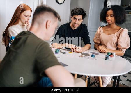 Gruppo di giovani amici multietnici che hanno festa a casa durante i fine settimana giocando a poker gioco di carte in soggiorno e parlando. Foto Stock