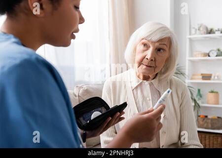 infermiera multirazziale che tiene la penna di lancet e che spiega come usarla alla donna anziana con i capelli grigi Foto Stock