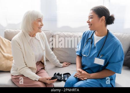 donna anziana felice che tiene glucometro vicino infermiere multirazziale in uniforme blu Foto Stock