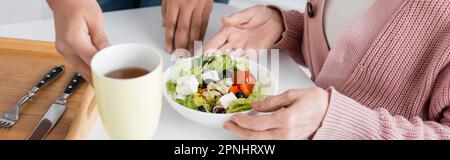vista ritagliata di un assistente sociale multirazziale che serve il pranzo vicino a una donna anziana, banner Foto Stock