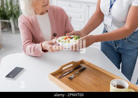 vista ritagliata di un assistente sociale multirazziale che dà ciotola con insalata a una donna anziana felice Foto Stock