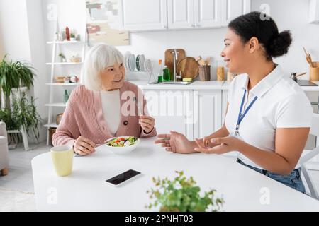 donna anziana felice con i capelli grigi che mangiano l'insalata e che parlano con l'assistente sociale multirazziale Foto Stock