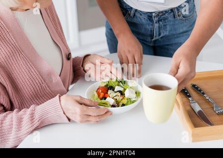 vista ritagliata di un assistente sociale multirazziale che serve il pranzo vicino a una donna anziana Foto Stock