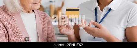vista ritagliata del lavoratore sociale multirazziale in possesso di carta di credito vicino a una donna anziana a casa, banner Foto Stock