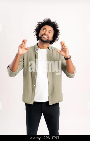 Vi auguriamo buona fortuna. Bel 30s curly ragazzo in camicia casual tenendo le dita incrociate isolato su bianco, carismatico afro-americano uomo sperando di miracolo, forte desiderio di qualcosa, in preghiera Foto Stock