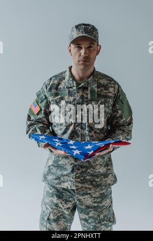 Soldato americano in uniforme che tiene piegata bandiera degli Stati Uniti durante la giornata commemorativa isolato sul grigio Foto Stock