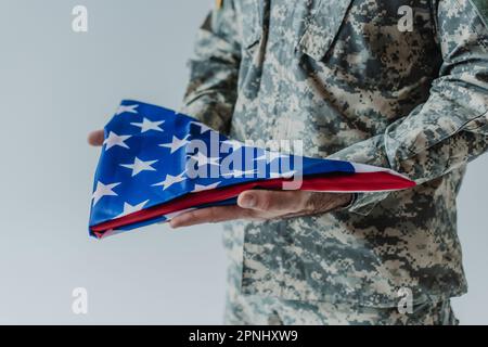 Vista parziale del soldato americano in uniforme che tiene la bandiera piegata degli Stati Uniti durante il giorno del memoriale isolato sul grigio Foto Stock