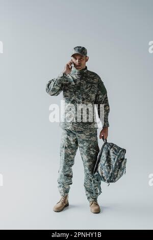 addetto alla manutenzione in berretto e uniforme militare che parla sullo smartphone mentre tiene lo zaino in grigio Foto Stock