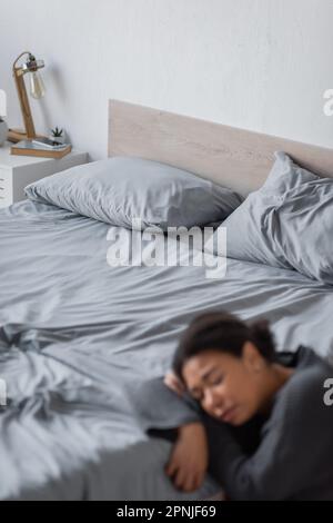 Donna multirazziale sfocata con depressione seduta vicino a letto a casa, immagine di scorta Foto Stock