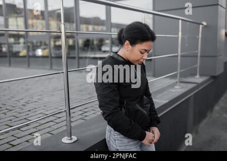 Giovane donna multirazziale con apatia in piedi vicino edificio all'aperto, immagine stock Foto Stock