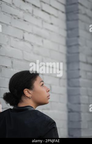 Vista laterale di donna multirazziale con problema psicologico in piedi vicino al muro di mattoni, immagine stock Foto Stock