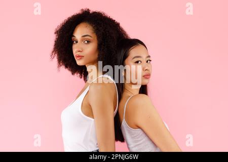 Ritratto di giovani belle donne asiatiche e afro-americane che si levano in piedi indietro, posate insieme su sfondo rosa Foto Stock