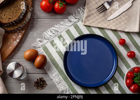 Una piastra blu vuota come servire sul tavolo. Tavolo con verdure e pane su sfondo grigio. Foto Stock