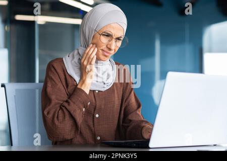 Primo piano. Mal di denti sul posto di lavoro. Ritratto di una giovane donna che lavora in ufficio a un tavolo con un computer portatile, tenendo la guancia, facendo un massaggio, sentendo un forte mal di denti. Foto Stock