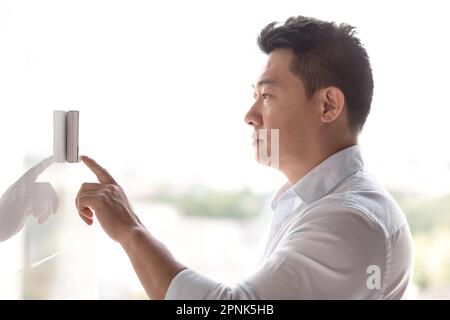 Un uomo asiatico si trova vicino a un pannello elettronico sul muro e preme con il dito la campana di ingresso di una porta automatica. Configura il condizionatore da parete, il sistema di sicurezza. Foto Stock