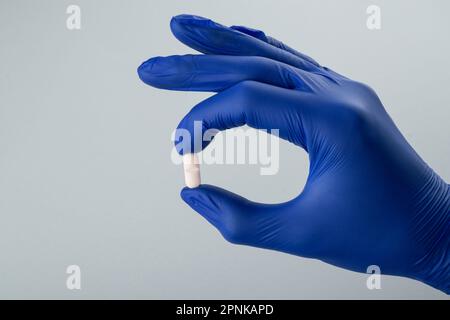 Mano maschile in un guanto medico blu che tiene una pillola bianca su uno sfondo grigio. Primo piano Foto Stock