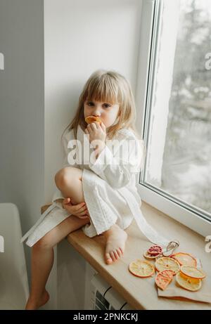 Una ragazza in un cappotto bianco è triste seduta sul davanzale, mangiando il marshmallow della frutta. Foto Stock